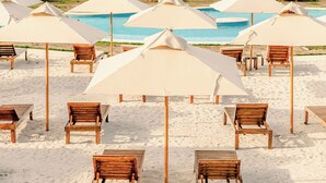 A beach with rows of wooden lounge chairs and beige umbrellas, a city skyline in the background, and a calm sea. - Anaho Beach Club (Cartagena)