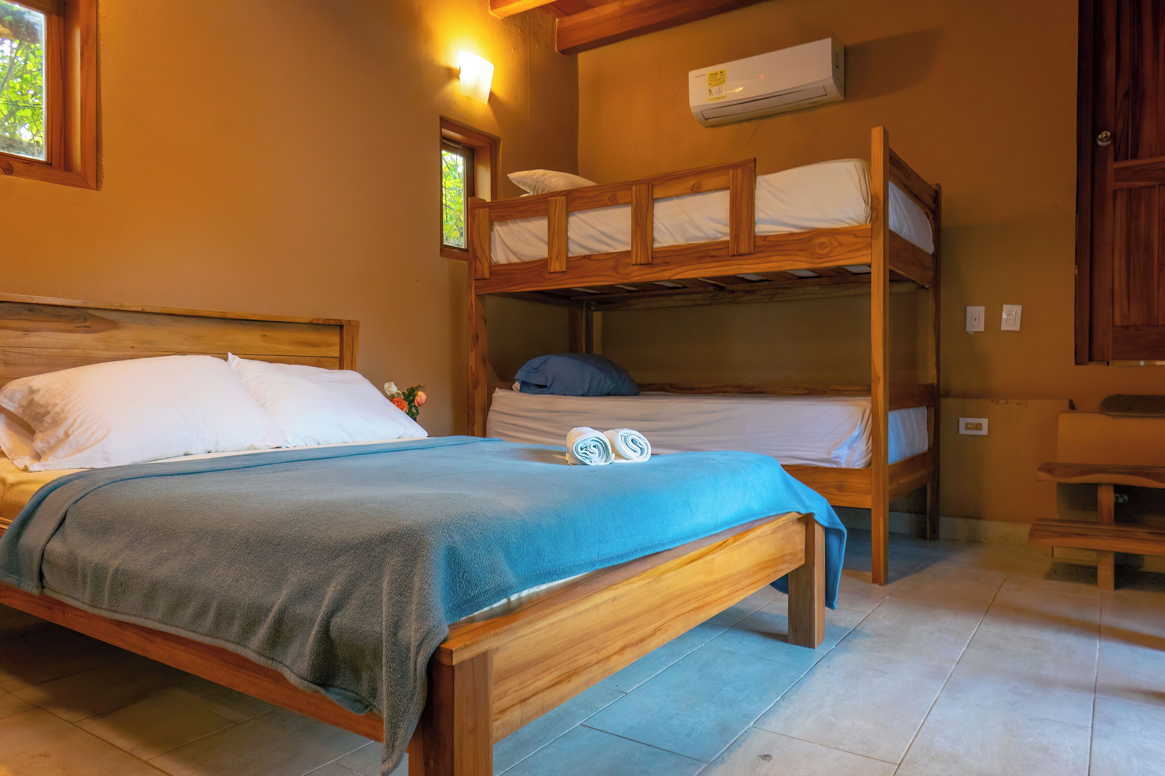 Une chambre avec un lit superposé et un lit simple. Il y a une fenêtre donnant sur un espace verdoyant, un climatiseur fixé au mur et un banc en bois.