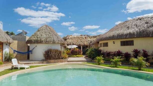 A pool area with thatched-roof buildings, a hammock, and a clear blue sky. - Casa con Habitación Doble, Casa Maya (Izamal)