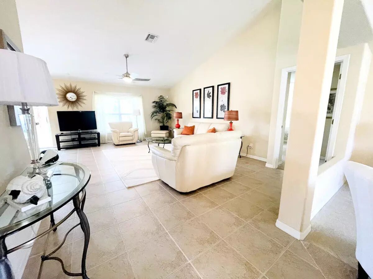 A spacious living room with a white sofa, a glass coffee table, and a TV. There is a ceiling fan and a large window providing natural light.