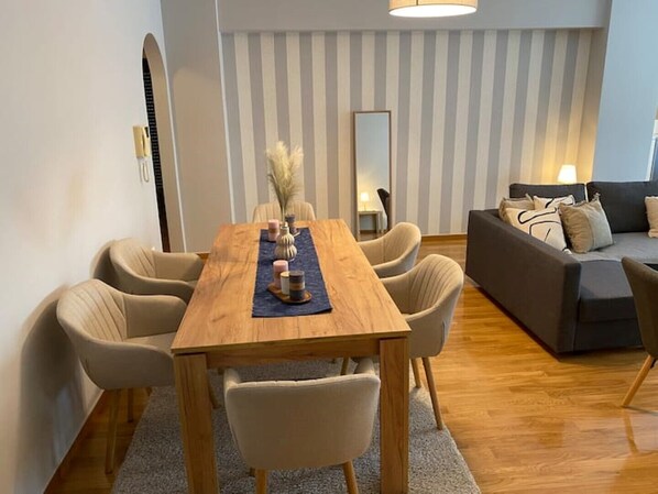 Uma sala de estar moderna com uma mesa de jantar de madeira, cadeiras bege, um sofá cinza e um espelho na parede.