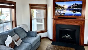 Een woonkamer met een open haard, een aan een houten wand bevestigde televisie en een bank met kussens.
