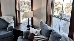 Een hoekkamer met een grijze bank, een kleine houten tafel, een lamp en een raam met uitzicht op woonhuizen.