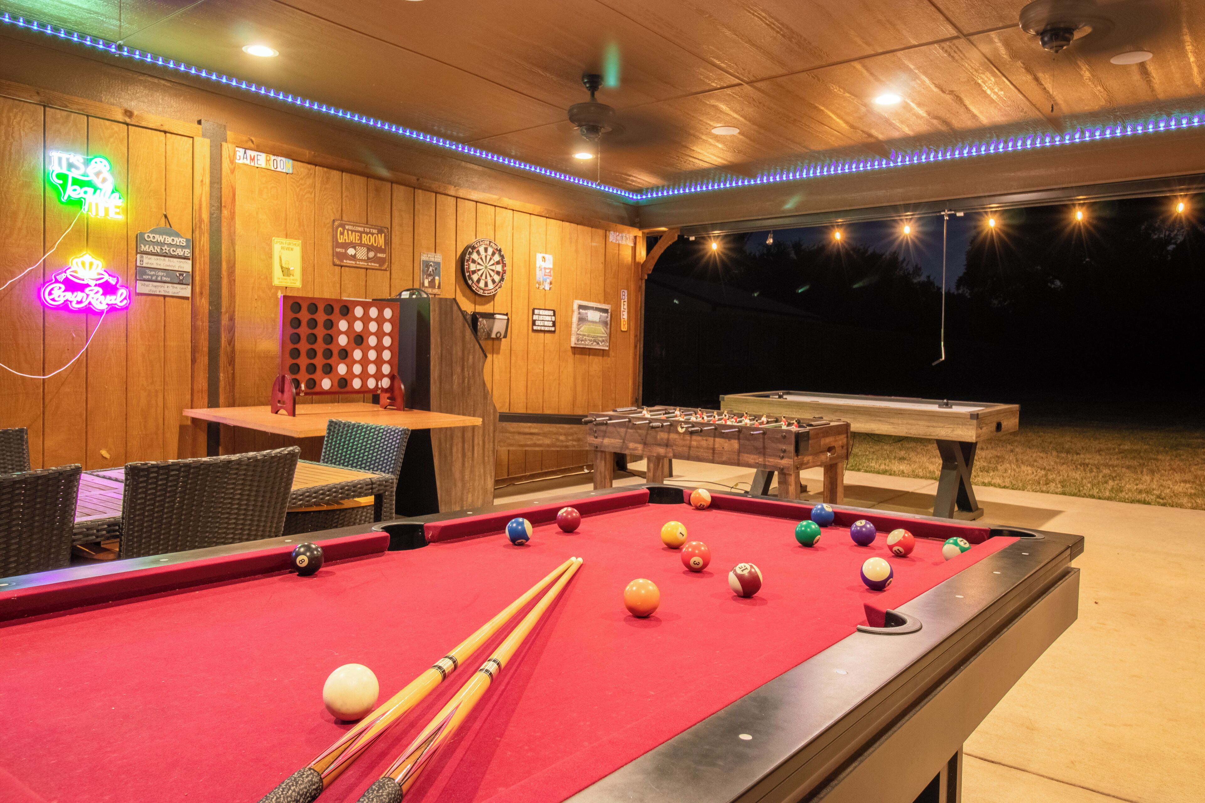 A pool table with billiard balls and cues, a dartboard, and a foosball table in a room with wooden walls and neon signs.