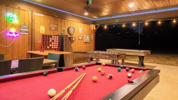 A pool table with billiard balls and cues, a dartboard, and a foosball table in a room with wooden walls and neon signs.