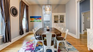Une salle à manger avec une table en bois, des chaises blanches, un tapis coloré et un tableau accroché au mur.
