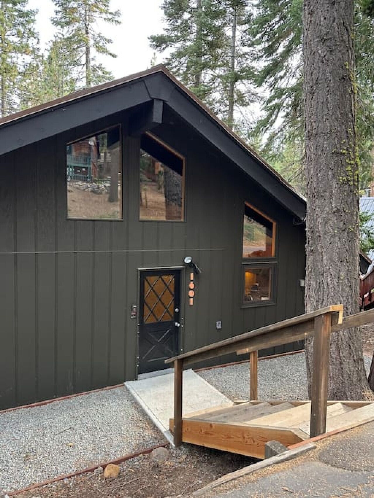 Une cabane aux couleurs sombres, dotée d’un escalier en bois menant à une porte, et entourée d’arbres.