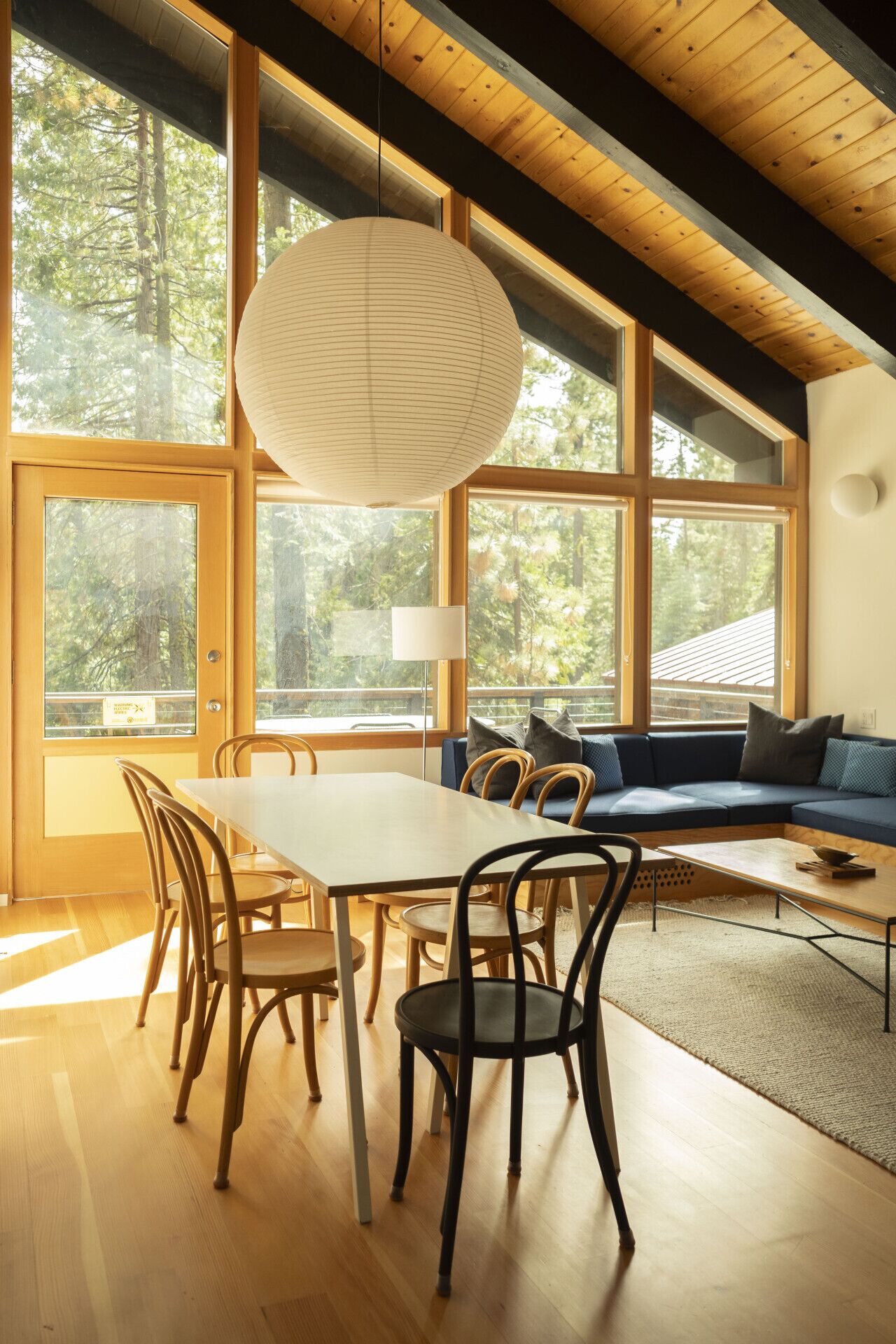 A modern interior with a dining table, chairs, a sofa, and large windows offering a view of the forest.