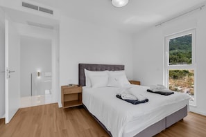 Une chambre moderne avec un grand lit, un plancher en bois, une tête de lit blanche et une vue sur un paysage montagneux par une grande fenêtre.