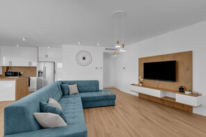 Uma sala de estar moderna com um sofá seccional, uma TV de tela plana fixada em uma parede de madeira, e uma cozinha ao fundo.