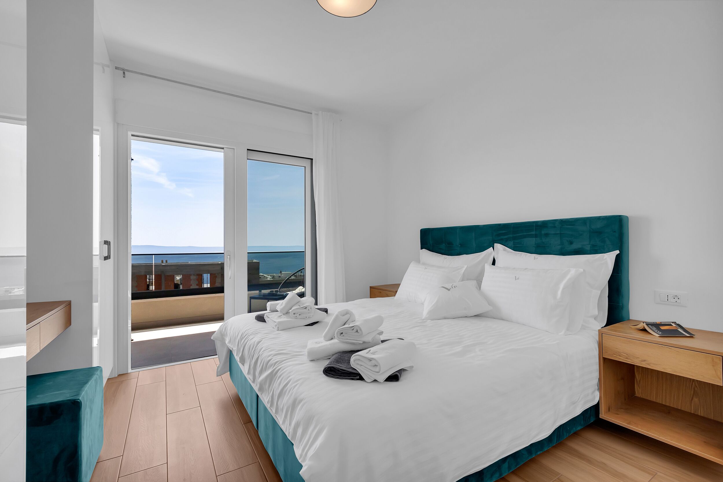 Une chambre d’hôtel moderne avec un grand lit, une table de chevet en bois, une vue sur la mer depuis un balcon et une atmosphère lumineuse et aérée.