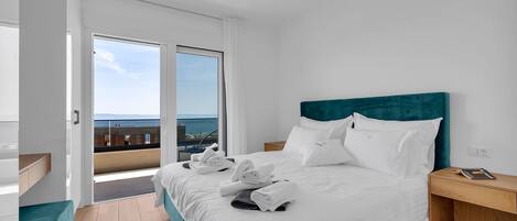 Habitación de hotel moderna con una cama grande, una mesita de noche de madera, vista al mar desde un balcón y un ambiente luminoso y espacioso.