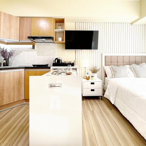 Una habitación de hotel moderna con área de cocina, televisor de pantalla plana montado en la pared, cama con ropa blanca y una mesita de noche con lámpara y objetos decorativos.
