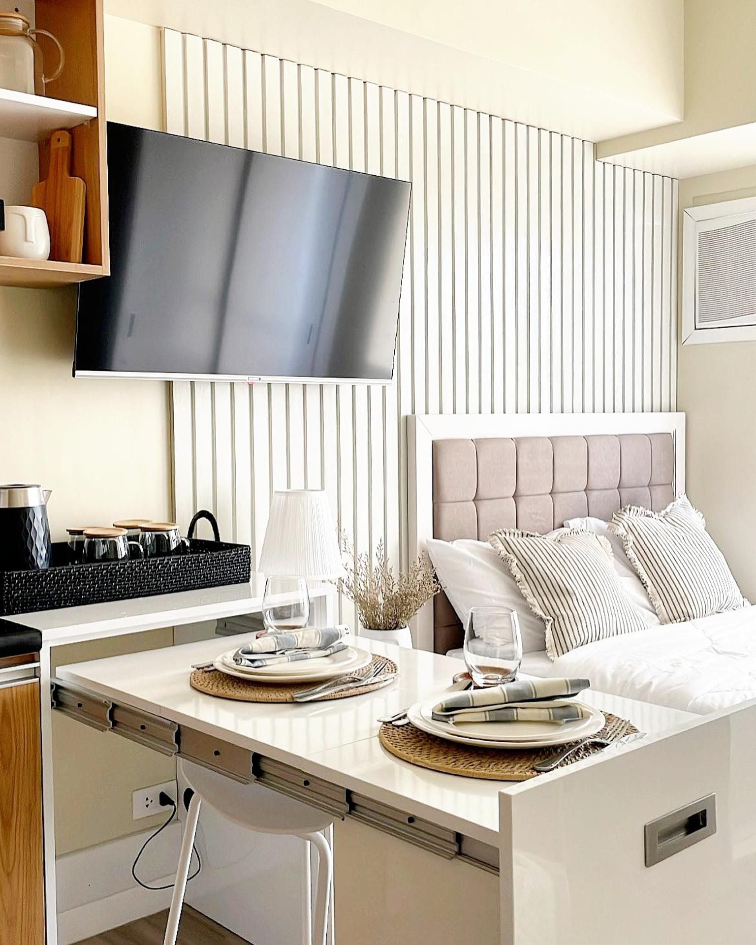 A modern hotel room with a flat-screen TV mounted on the wall, a bed with a tufted headboard, and a dining area with a table set for two.