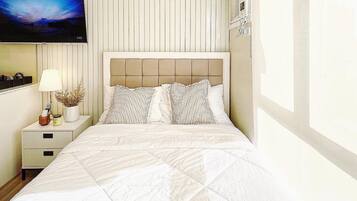 Quarto com cama, mesa de cabeceira, abajur, televisão e janela.