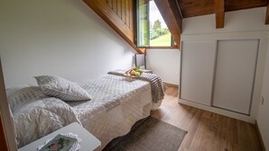 Une chambre avec un plafond mansardé, un lit recouvert d’une courtepointe, une table de chevet agrémentée d’un objet décoratif et une fenêtre donnant sur un paysage verdoyant.