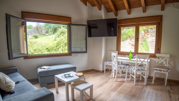 Un salon confortable avec un canapé, une petite table et un coin repas avec des chaises. De grandes fenêtres offrent une vue sur la verdure environnante.