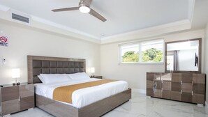A modern bedroom with a bed, nightstands, a dresser, a ceiling fan, and a window with a view of greenery.