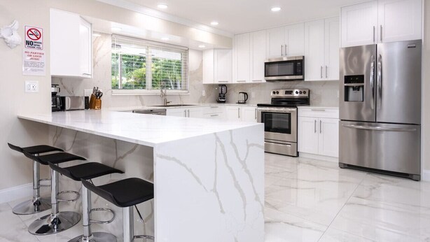 A modern kitchen with white marble countertops, stainless steel appliances, and a bar area with stools.