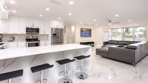 A modern kitchen with white cabinets, stainless steel appliances, a marble island with bar stools, and a grey sofa in a bright, well-lit room with large windows.
