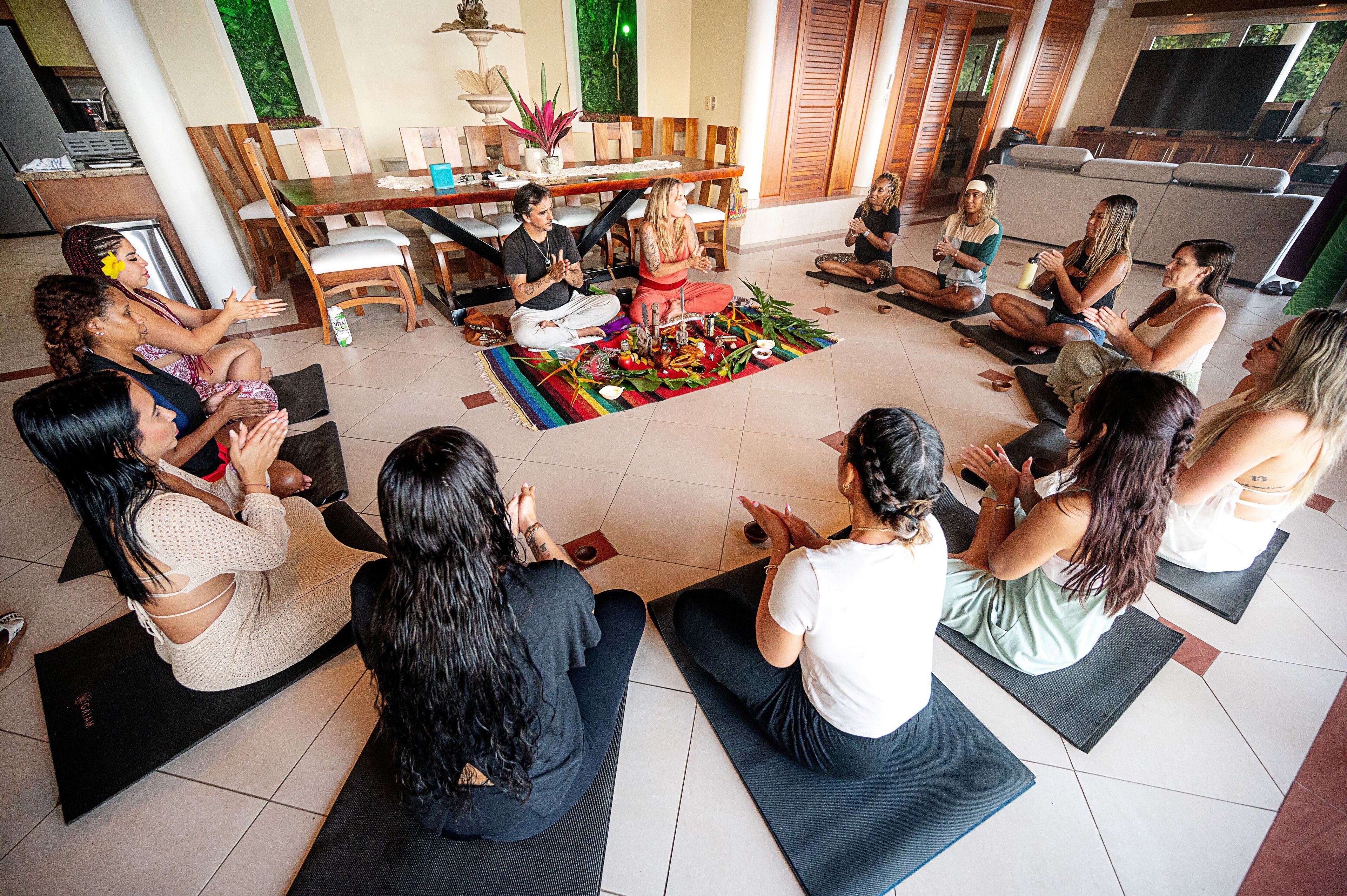 A group of people sitting on yoga mats in a room with a dining table and chairs, a TV, and a plant.