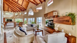 A spacious living area with a high ceiling, large windows, and a modern kitchen.