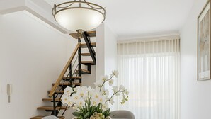Un comedor moderno con una mesa de cristal, cuatro sillas, un jarrón de orquídeas blancas, una lámpara colgante y una escalera al fondo.