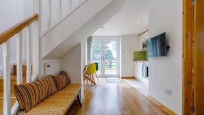 A spacious living area with a wooden staircase, a bench with patterned cushions, a bright yellow chair, and a kitchen with a flat-screen TV. - Charming 2-bed Cottage, Beautiful Views, Knutsford (Knutsford)