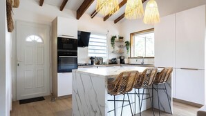 A modern kitchen with a marble island, black appliances, wicker bar stools, and pendant lighting. - Charmante Villa Neuve à Trinité (La Trinité)