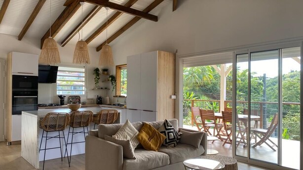A modern kitchen with a central island, bar stools, and a dining area with wooden chairs. - Charmante Villa Neuve à Trinité (La Trinité)