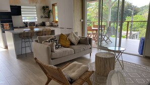 A modern living space with a sofa, wicker chairs, a dining area, and a view of a lush garden through large sliding doors. - Charmante Villa Neuve à Trinité (La Trinité)