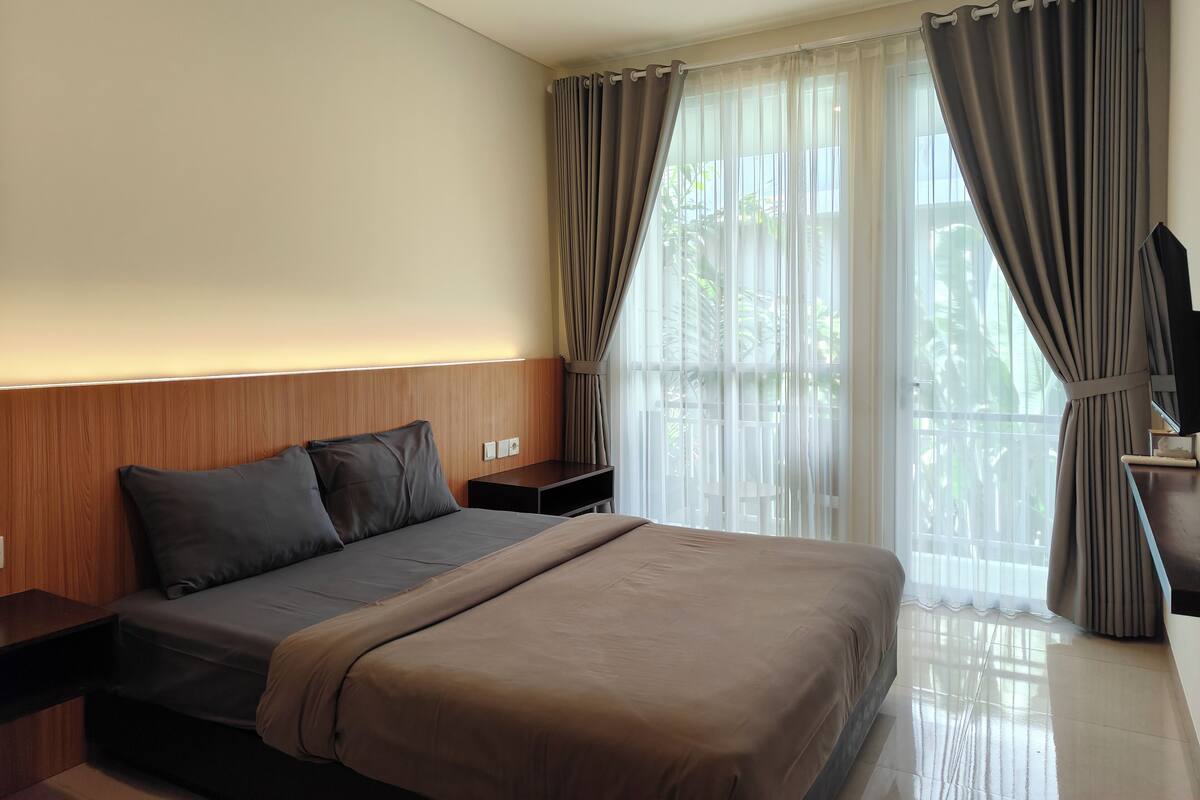 A modern hotel room with a large bed, a wooden headboard, a bedside table, and a TV.