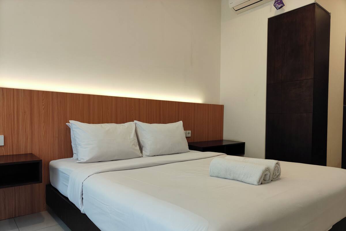 A neatly made bed with white linens and pillows, a wooden headboard, a dark wooden wardrobe, and a small bedside table with a lamp.