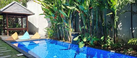 A swimming pool with a tiled edge, surrounded by lush green plants and a small gazebo.