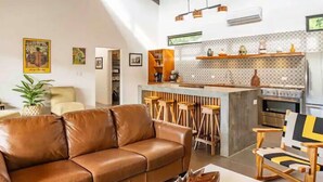 A modern living room with a brown leather sofa, a wooden bar with stools, and a kitchen area with patterned backsplash and stainless steel appliances. - Casa Tranquila, Brand NEW Modern Villa with Pool (Sámara)