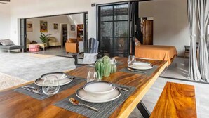 A modern dining area with a wooden table set for four, a large window, and a view of the living room. - Casa Tranquila, Brand NEW Modern Villa with Pool (Sámara)