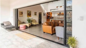 A modern living room with a sofa, armchairs, and a coffee table. There is a sliding glass door leading to a kitchen area with shelves and a countertop. - Casa Tranquila, Brand NEW Modern Villa with Pool (Sámara)