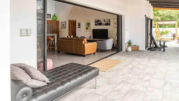 A modern living room with a sofa, a television, and a dining area visible through an open door. - Casa Tranquila, Brand NEW Modern Villa with Pool (Sámara)