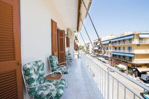 A balcony with a row of tropical-themed chairs and a small table, overlooking a street with buildings and shops.