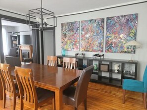 A dining area with a wooden table and chairs, a black metal ceiling light, and a wall with abstract paintings.