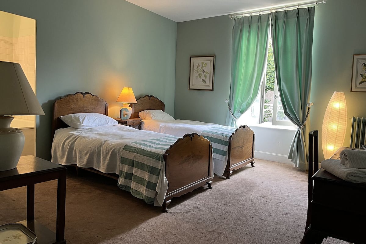 A bedroom with two wooden beds, a nightstand with a lamp, a window with green curtains, and a bedside table with a lamp.