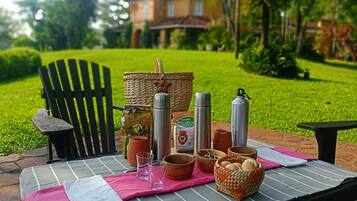 Un montaje para picnic con una mesa, sillas y diversos alimentos y bebidas en un área cubierta de césped, con una casa al fondo.