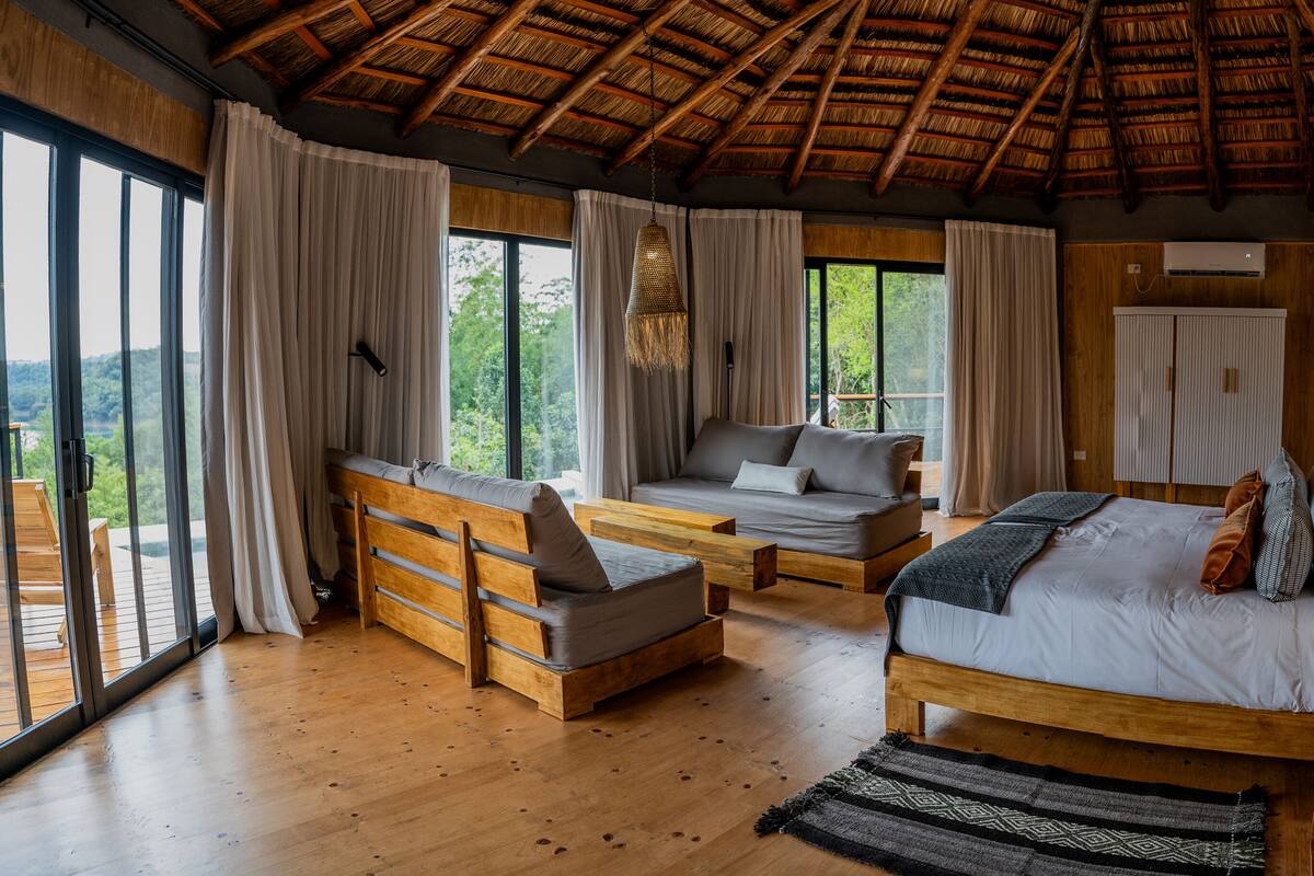 Une chambre spacieuse avec un grand lit, un canapé et une banquette, le tout en bois. De grandes fenêtres sont agrémentées de rideaux, le toit est fait de chaume et un climatiseur est fixé au mur.