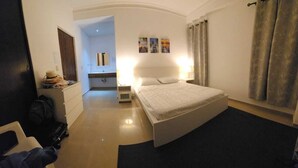 Ein Hotelzimmer mit einem Bett, einem Schreibtisch mit Lampe, einem Kleiderschrank und einem durch einen Bogen sichtbaren Badezimmer.