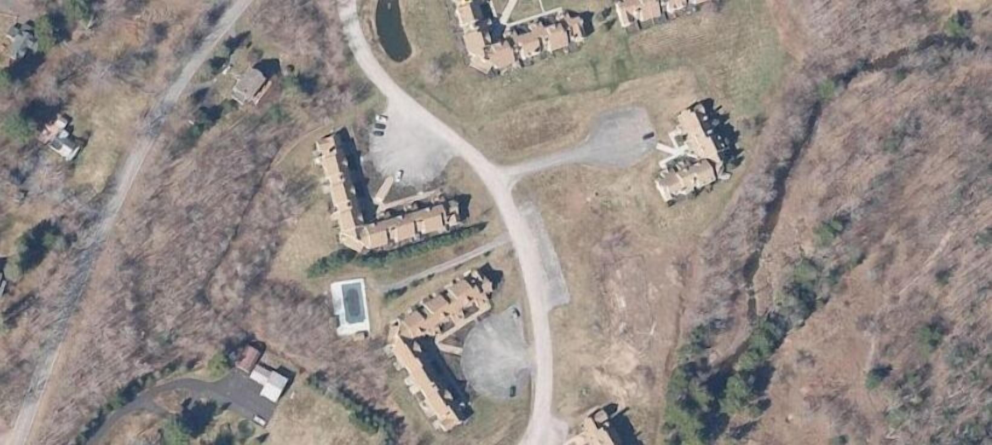 An aerial view of a residential area with multiple houses, a central road, and a small body of water.