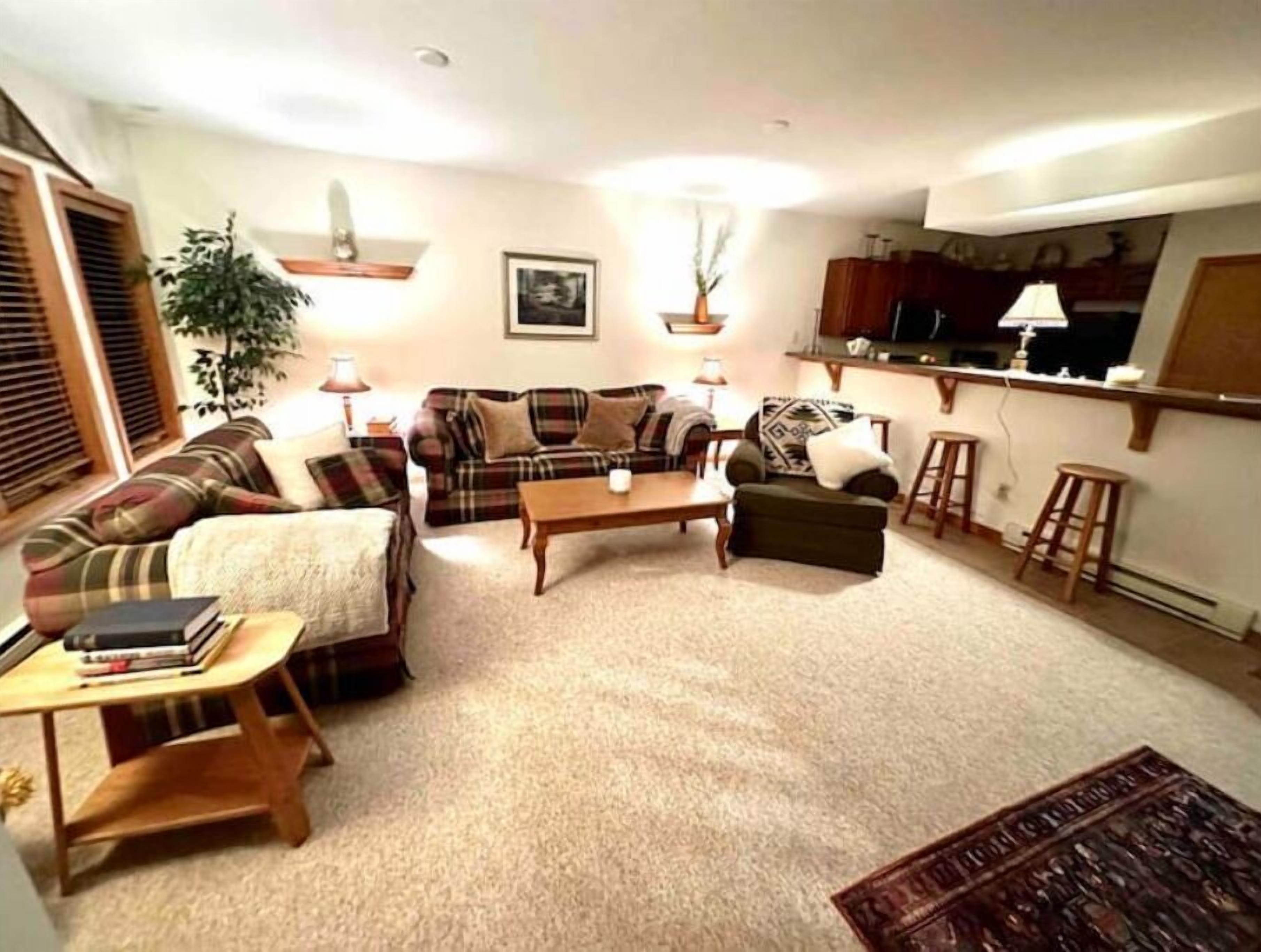 A cozy living room with a sofa, a coffee table, and a small table with books. There is a plant, a picture on the wall, and a kitchen area in the background.