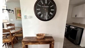 A dining area with a wooden table and chairs, a wall-mounted clock, and a kitchen in the background.