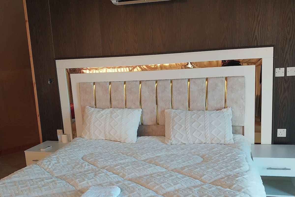 Habitación de hotel con una cama grande, cabecera con líneas verticales, unidad de aire acondicionado blanca sobre la cama y una mesita de noche con una lámpara.