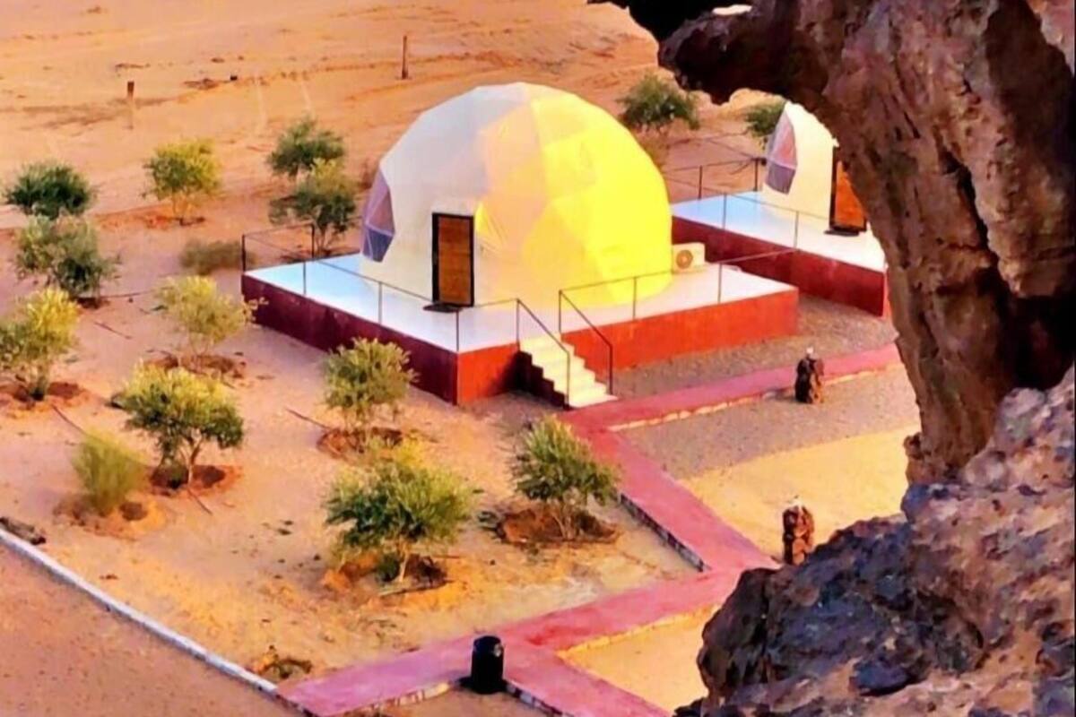 Paisaje desértico con una estructura esférica distintiva, rodeada de pequeños árboles y un afloramiento rocoso.