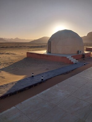 A dome-shaped building with a door and steps leading up to it, situated in a desert landscape with mountains in the background. - eisam camp (Wadi Rum)